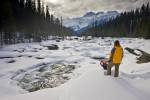 A beautiful scene on the banks of the snowcovered Mistaya River during winter in Banff National Park, Alberta, Canada. Mounta Sarbach towers above the forest in the background while a tourist takes in the scenery.
