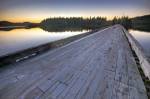 An old wood bridge leads towards the tree lined shores of Kennedy Lake on Vancouver Island. The trees are silhouetted by the sunset on a beautiful calm evening.